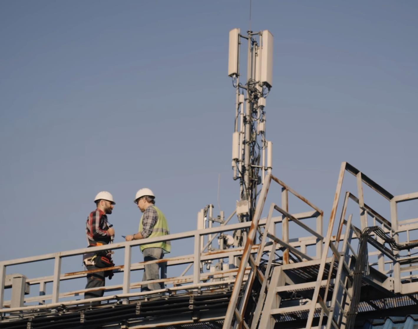 Two field workers in hard hats consulting on a rooftop next to a cell tower installation